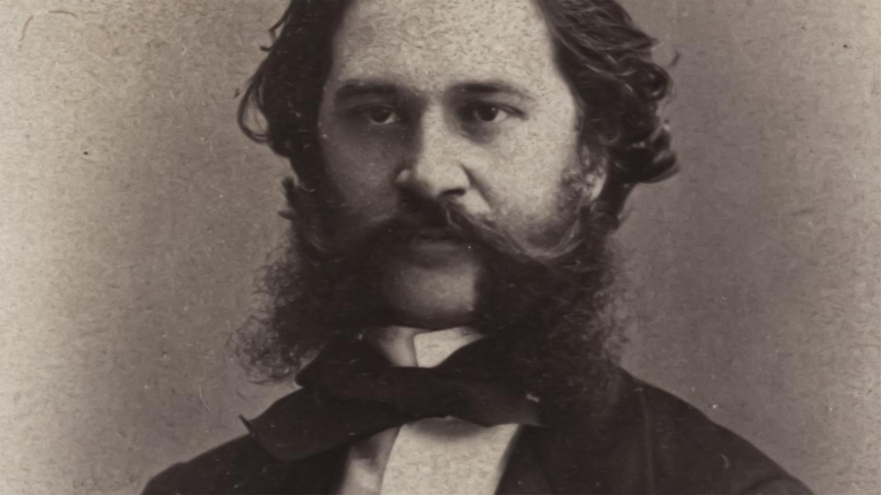 Das Bild zeigt ein Porträt eines Mannes in jungen Jahren, der Johann Strauss darstellt. Er hat einen vollen, dunklen Bart und eine ähnliche Frisur, die zu den modischen Stilen des 19. Jahrhunderts passt. Seine Augen sind direkt auf den Betrachter gerichtet, und er trägt einen eleganten Anzug mit einem weißen Hemd und einer schwarzen Fliege. Der Hintergrund des Bildes ist unscharf und neutral gehalten, was den Fokus auf das Gesicht des Porträtierten lenkt. Die fotografische Technik vermittelt einen historischen Eindruck, der typisch für die damalige Zeit ist.