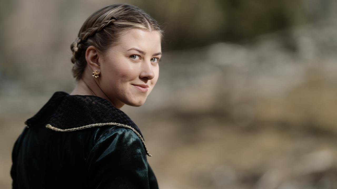 Das Bild zeigt eine Frau mit einem lächelnden Gesichtsausdruck, die leicht zur Seite gedreht ist. Sie trägt ein elegantes, dunkles Kleid, das einen schwarzen Kragen mit goldenen Verzierungen hat. Ihre Haare sind zu einem geflochtenen Zopf gestylt, und sie trägt kleine, runde Ohrringe. Der Hintergrund ist unscharf, scheint aber einen natürlichen, möglicherweise ländlichen oder historisch angehauchten Kontext zu haben, da die Farben eher warm und erdig sind.   Das Bild gehört zu einem Filmprojekt mit dem Titel "Der Sagenjäger", in dem es um eine dramatische Geschichte im Mittelalter geht, die Themen wie Liebe, Eifersucht und Tod behandelt.