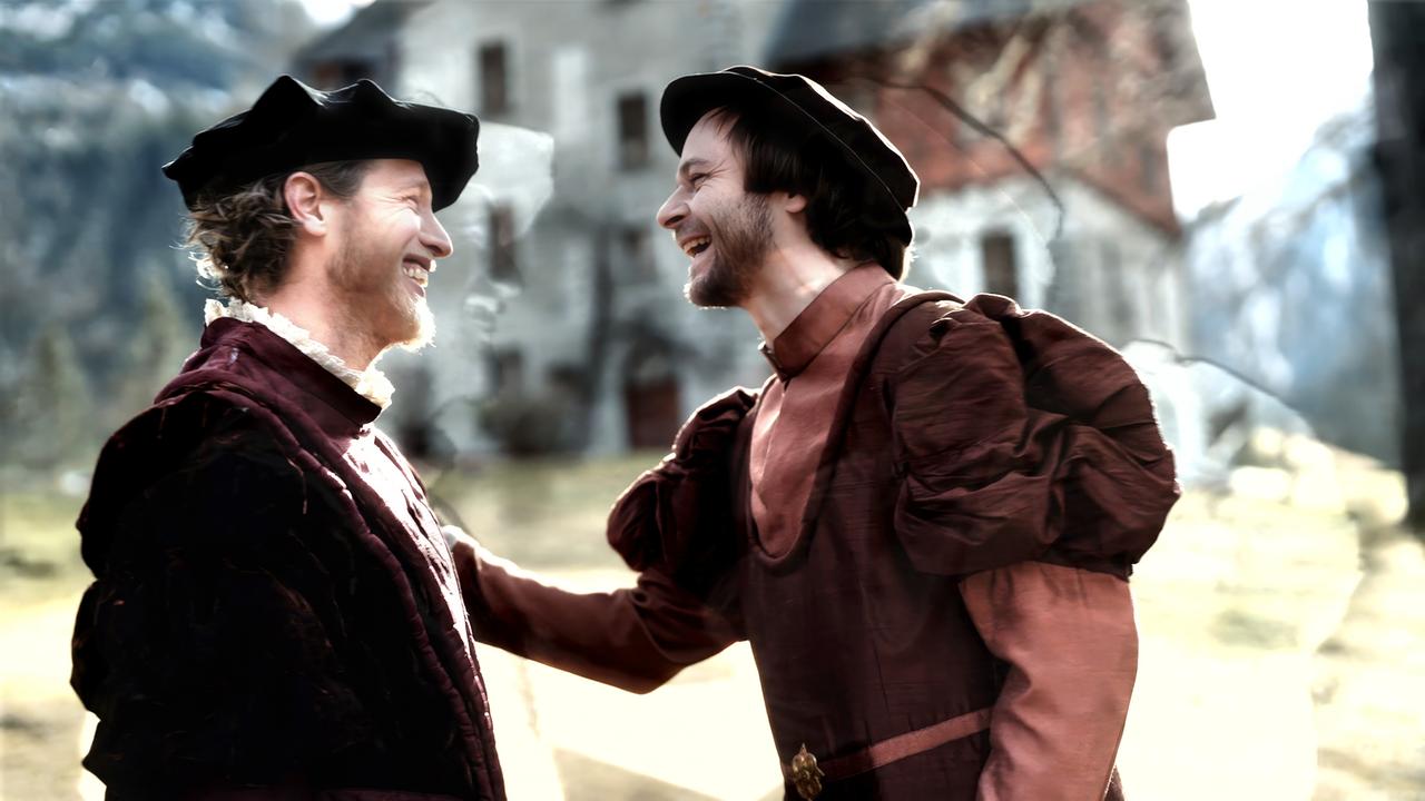 Das Bild zeigt zwei Männer, die sich herzlich miteinander unterhalten und Lächeln austauschen. Beide tragen historische Kostüme, die an das Mittelalter erinnern. Der Mann auf der linken Seite trägt ein burgunderfarbenes Gewand mit einem weißen, rüschierten Kragen, während der Mann auf der rechten Seite ein braunes Gewand mit weit ausgestellten Ärmeln und einem ähnlichen Kragen trägt. Beide haben Hüte auf, die ebenfalls dem Stil der Zeit entsprechen.  Im Hintergrund ist ein historisches Gebäude zu sehen, das rustikal wirkt und eine Mischung aus Stein und Holz darstellt. Es scheint sich in einer ländlichen Umgebung zu befinden, mit Bergen und Bäumen im Hintergrund. Die Szenerie wirkt freundlich und einladend, und die Stimmung zwischen den beiden Männern scheint positiv und aufgeschlossen zu sein.