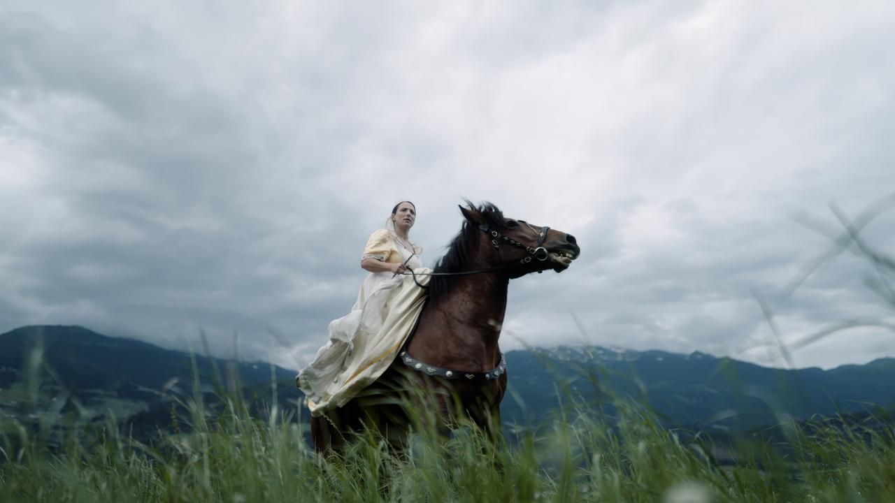Das Bild zeigt eine Frau, die auf einem braunen Pferd sitzt. Sie trägt ein elegantes, hellbeiges Kleid mit schulterfreien Ärmeln, das bis zum Boden reicht. Die Frau hat ihre Haare locker frisiert und schaut ernst in die Ferne. Im Hintergrund erstrecken sich grüne Wiesen und Berge, die teilweise von Wolken bedeckt sind. Der Himmel ist bewölkt und vermittelt eine düstere Stimmung. Das Bild scheint aus einer Filmszene zu stammen, die möglicherweise in einer historischen oder fantastischen Umgebung spielt. Es gibt keine sichtbaren Gebäude oder spezifische Orte im Hintergrund, nur die natürliche Landschaft.