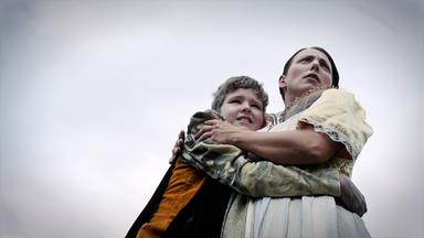 Das Bild zeigt zwei verkleidete Personen, die offensichtlich eine Reenactment-Szene darstellen. Auf der linken Seite ist ein Kind zu sehen, das in einem grauen und braunen Outfit gekleidet ist. Es umarmt eine erwachsene Frau, die zur rechten Seite des Bildes steht. Diese Frau trägt ein helles, historisch anmutendes Kleid mit einer auffälligen Kette um den Hals. Beide Charaktere zeigen besorgte Gesichtsausdrücke und blicken nach oben, als würden sie etwas beobachten oder fürchten. Der Hintergrund ist wolkig und wirkt düster, was zur dramatischen Stimmung der Szene beiträgt. Die Umgebung ist nicht klar definiert, aber es vermittelt den Eindruck einer natürlichen Kulisse.