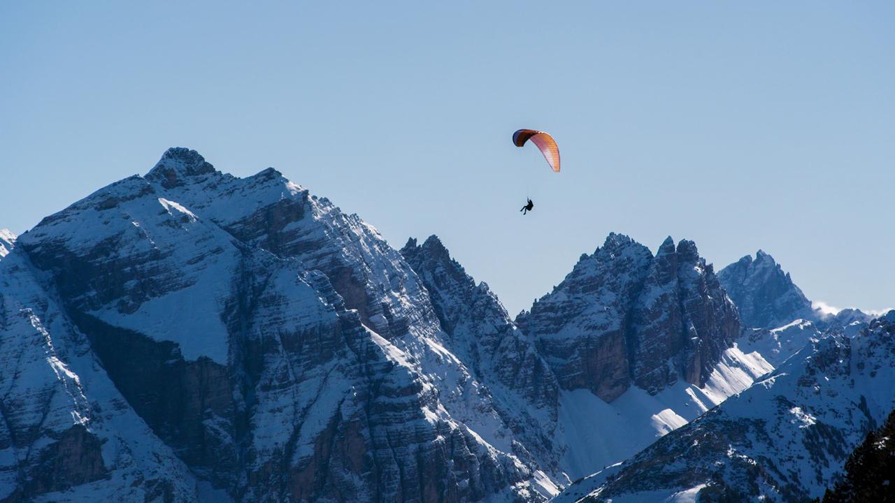Ein Paragleiter schwebt über schneebedeckte Berge. Die Gipfel sind steil und rau, der Himmel ist klar und blau. Die Szenerie vermittelt eine alpine Landschaft.
