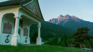 Im Bild ist eine Kapelle mit dekorativem Dach und Säulen zu sehen. Sie steht vor einer Berglandschaft. Im Hintergrund zeigen sich hohe, mit Schnee bedeckte Berge und bewaldete Hänge.