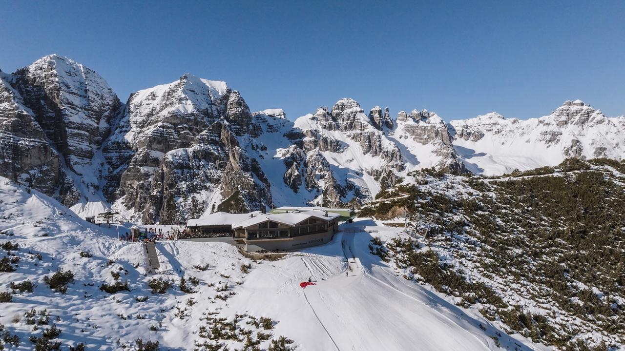 Die Aufnahme zeigt hohe, schneebedeckte Berge unter einem blauen Himmel. Im Vordergrund liegt ein Gebäude, umgeben von Schnee und einer Skipiste. Menschen sind zu sehen, die sich im Freien aufhalten.