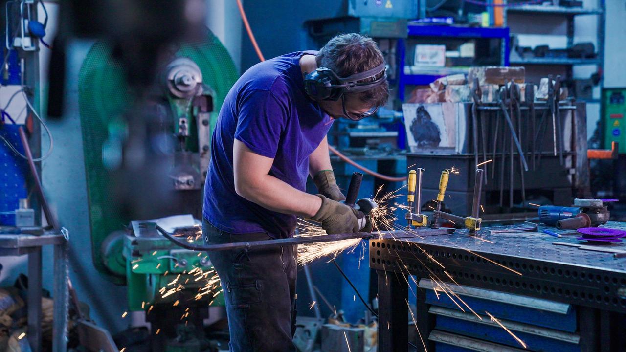 Ein Handwerker steht an einem Tisch in einer Werkstatt und arbeitet mit einer Schleifmaschine. Funken sprühen, während er ein Metallstück bearbeitet. Im Hintergrund sind Werkzeuge und Ausrüstung sichtbar.