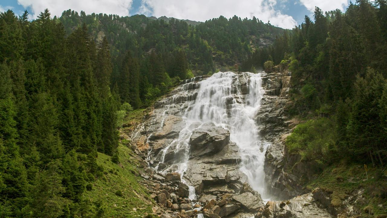 Im Bild ist der Grawa-Wasserfall zu sehen, der über felsige Stufen in ein Waldgebiet stürzt. Umgeben von grünen Bäumen und Felsen, fließt das Wasser mit viel Schwung. Der Himmel ist teils bewölkt.