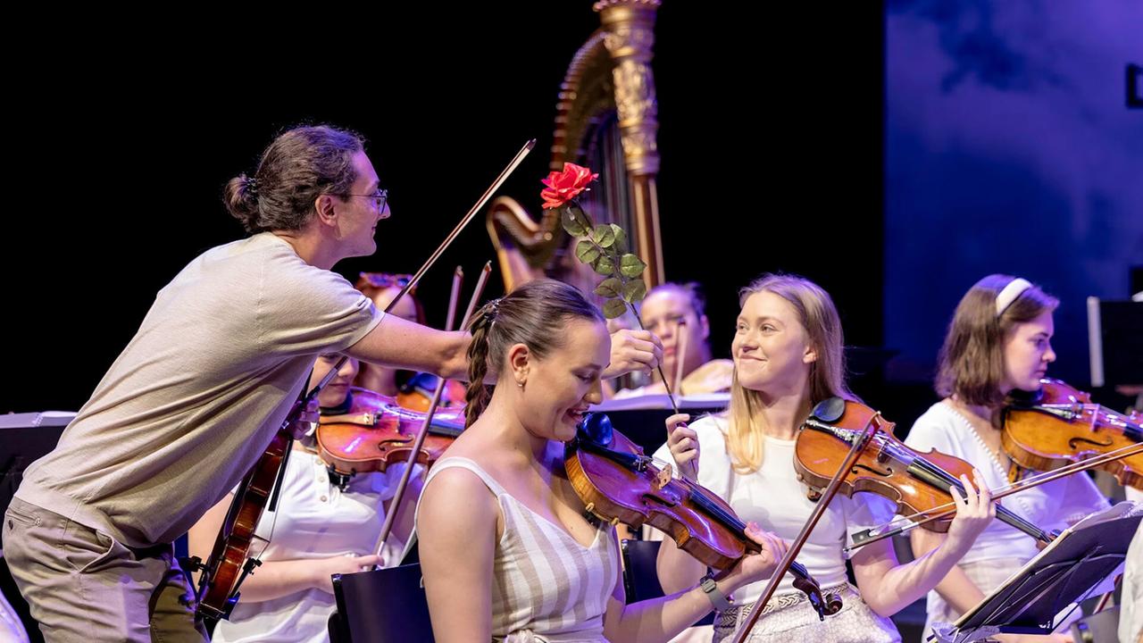 Das Bild zeigt eine Szene von einem Konzert des Styriarte Youth Orchestra. Im Vordergrund sind mehrere junge Musikerinnen zu sehen, die Geigen spielen. Eine Musikerin trägt ein gestreiftes Kleid und lächelt, während eine andere Musikerin in einem weißen Oberteil mit langen, blonden Haaren ebenfalls lächelt.   Ein männlicher Musiker steht in der Nähe und hält eine rote Rose in der Hand. Sein Gesicht ist teilweise zum Publikum gedreht. Im Hintergrund sind andere Orchestermitglieder sowie eine Harfe sichtbar. Der Bühnenhintergrund ist dunkel, was die Musiker und ihre Instrumente hervorhebt. Die Atmosphäre wirkt fröhlich und entspannt, möglicherweise während einer Pause oder in einer lebhaften Interaktion.   Die Musikerinnen und Musiker scheinen Teil eines Auftritts zu sein, der Gustavo Mahlers 2. Symphonie gewidmet ist.