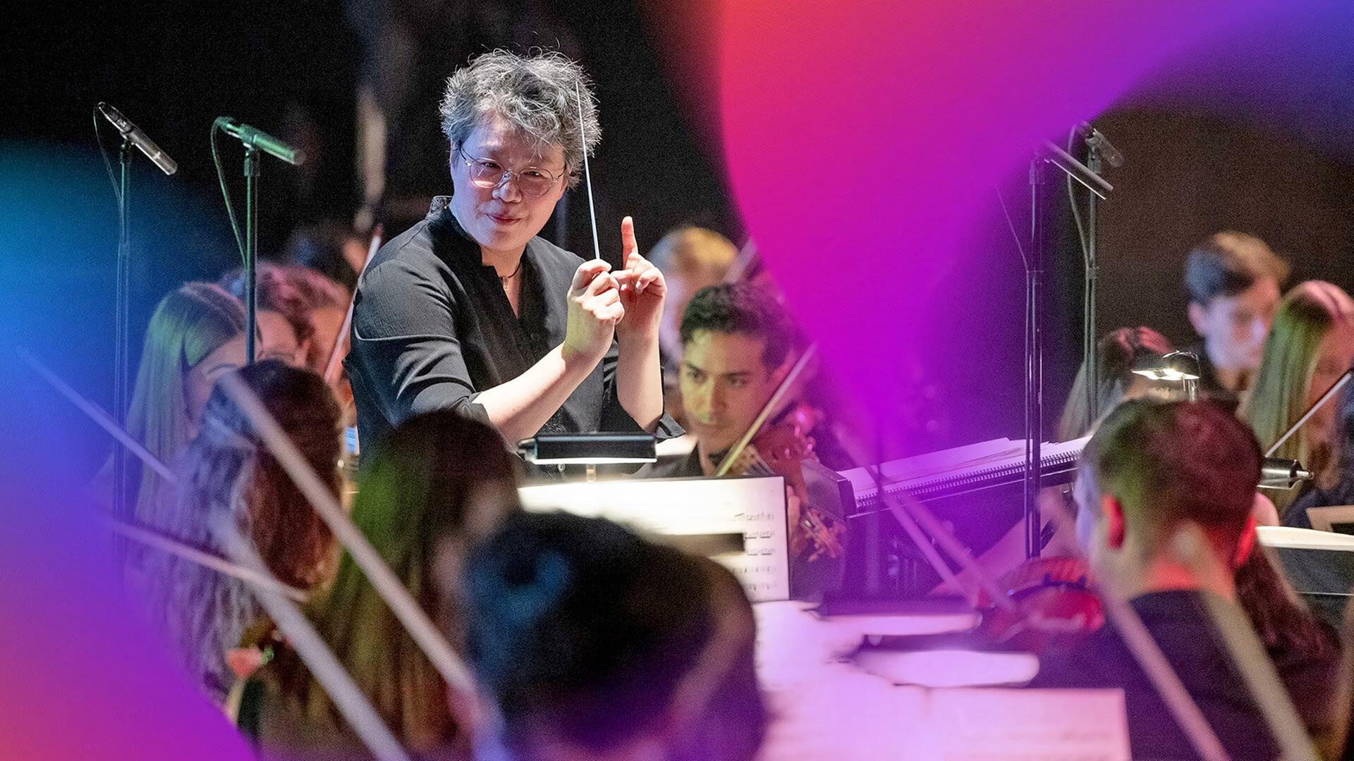 Auf dem Bild ist eine musikalische Aufführung zu sehen. Im Vordergrund steht eine Dirigentin mit kurzen, grauen Haaren, die eine schwarze Bluse trägt. Sie hält einen Dirigentenstab in einer Hand und gestikuliert mit der anderen Hand, während sie das Orchester leitet. Hinter ihr sind junge Musikerinnen und Musiker zu sehen, die Instrumente spielen.   Die Musiker sind teilweise in dunkler Kleidung gekleidet, und einige von ihnen sind mit ihren Instrumenten, wie Violinen, beschäftigt. Es gibt mehrere Mikrophonständer im Bild, und der Hintergrund ist leicht verschwommen, was den Fokus auf die Dirigentin und das Orchester lenkt.   Die Aufnahme vermittelt eine lebendige Atmosphäre des Konzerts, während die Dirigentin die Darbietung energetisch leitet. Es sind keine spezifischen Orte oder Gebäude im Bild sichtbar, lediglich die Auftrittssituation.