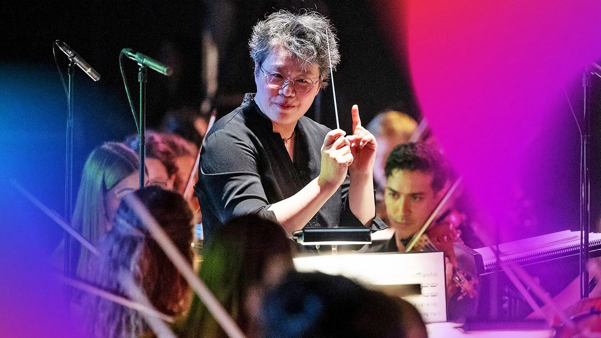 Auf dem Bild ist eine musikalische Aufführung zu sehen. Im Vordergrund steht eine Dirigentin mit kurzen, grauen Haaren, die eine schwarze Bluse trägt. Sie hält einen Dirigentenstab in einer Hand und gestikuliert mit der anderen Hand, während sie das Orchester leitet. Hinter ihr sind junge Musikerinnen und Musiker zu sehen, die Instrumente spielen.   Die Musiker sind teilweise in dunkler Kleidung gekleidet, und einige von ihnen sind mit ihren Instrumenten, wie Violinen, beschäftigt. Es gibt mehrere Mikrophonständer im Bild, und der Hintergrund ist leicht verschwommen, was den Fokus auf die Dirigentin und das Orchester lenkt.   Die Aufnahme vermittelt eine lebendige Atmosphäre des Konzerts, während die Dirigentin die Darbietung energetisch leitet. Es sind keine spezifischen Orte oder Gebäude im Bild sichtbar, lediglich die Auftrittssituation.
