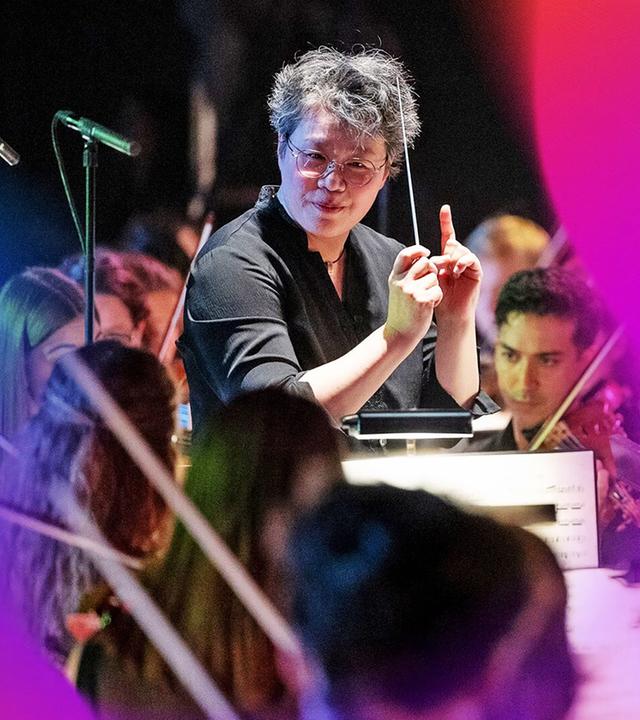 Auf dem Bild ist eine musikalische Aufführung zu sehen. Im Vordergrund steht eine Dirigentin mit kurzen, grauen Haaren, die eine schwarze Bluse trägt. Sie hält einen Dirigentenstab in einer Hand und gestikuliert mit der anderen Hand, während sie das Orchester leitet. Hinter ihr sind junge Musikerinnen und Musiker zu sehen, die Instrumente spielen.   Die Musiker sind teilweise in dunkler Kleidung gekleidet, und einige von ihnen sind mit ihren Instrumenten, wie Violinen, beschäftigt. Es gibt mehrere Mikrophonständer im Bild, und der Hintergrund ist leicht verschwommen, was den Fokus auf die Dirigentin und das Orchester lenkt.   Die Aufnahme vermittelt eine lebendige Atmosphäre des Konzerts, während die Dirigentin die Darbietung energetisch leitet. Es sind keine spezifischen Orte oder Gebäude im Bild sichtbar, lediglich die Auftrittssituation.