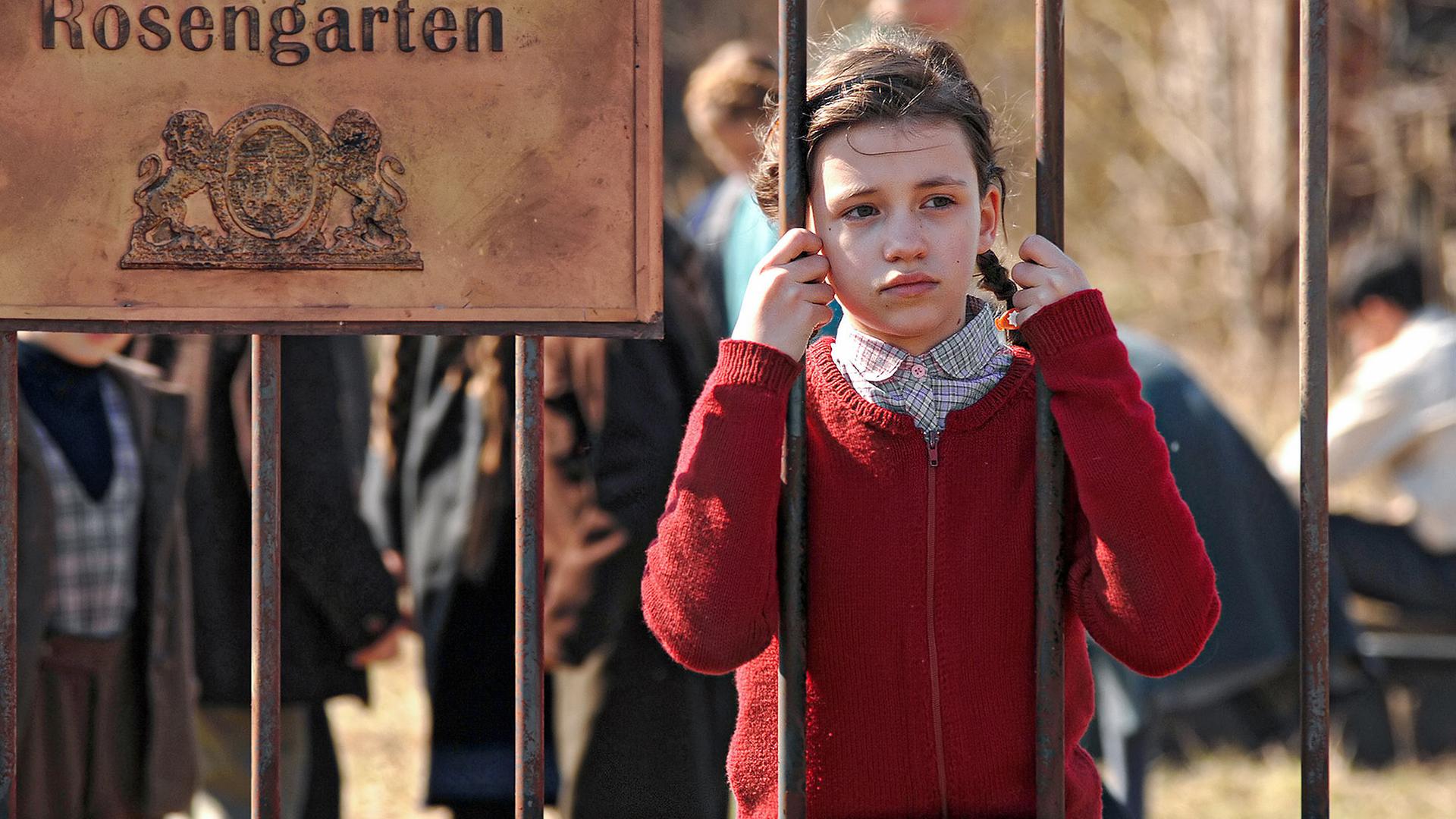Mädchen an Gitterstäben eines Zaunes, daneben ein Schild, auf dem steht: "Waisenhaus Rosengarten"  