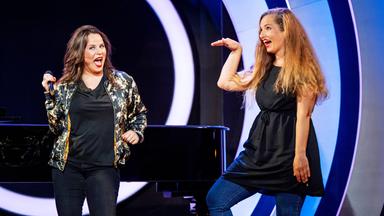 Julia Gamez Martin steht auf der linken Bildseite. Auf der rechten Bildseite ist Ariane Müller zu sehen. Beide haben den Mund geöffnet. Ariane Müller hebt ihre rechte Hand. Julia Gamez Martin stemmt ihre linke Hand in die Hüfte.