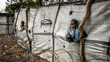 Kinder schauen aus Schlitzen in einer Plane am Zaun eines Flüchtlingslagers.