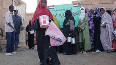 Verhüllte Frauen stehen mit Metallbehältern für Essen an.