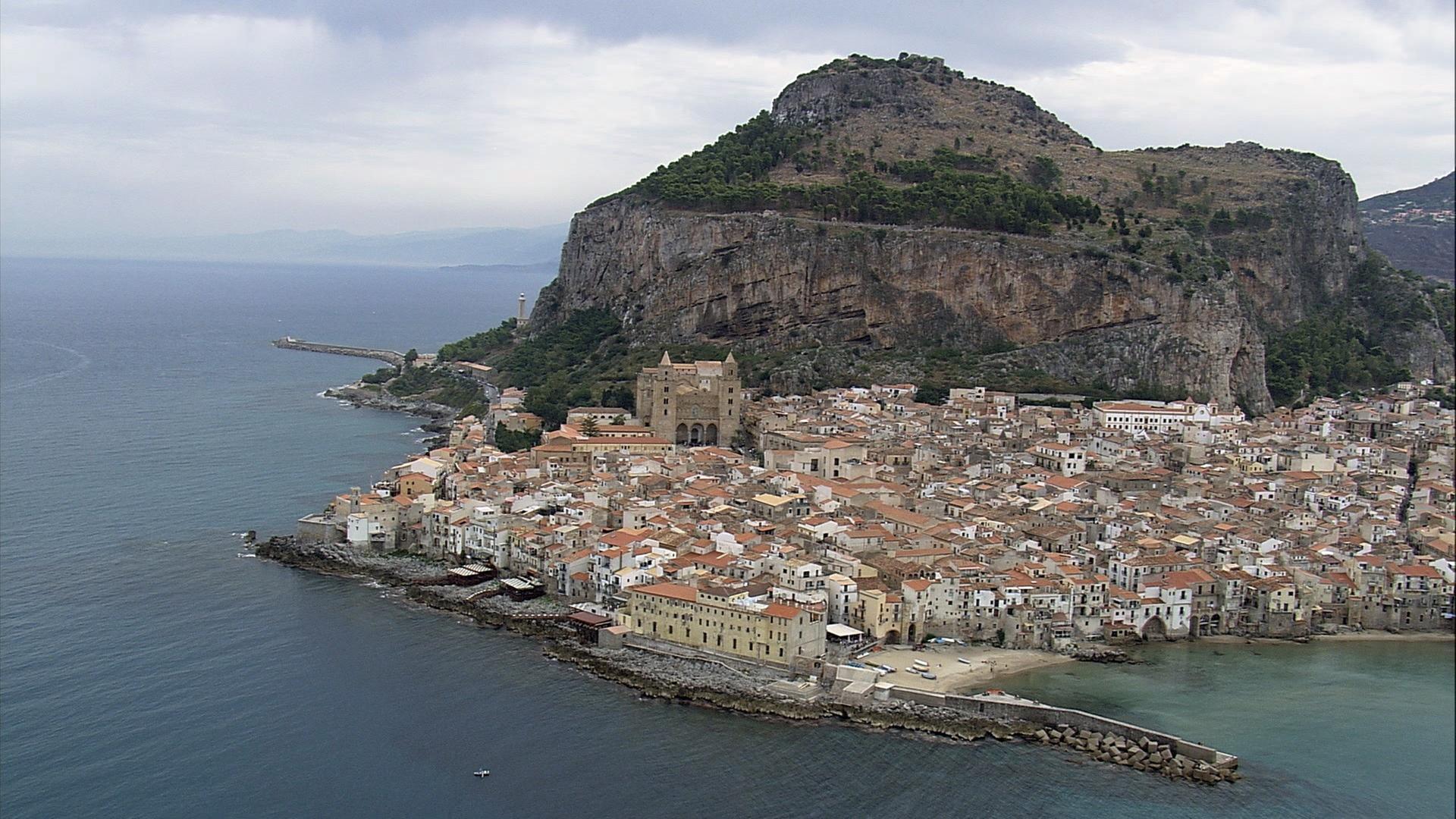 Die Küstenstadt Cefalù auf Sizilien mit großem Kirchengebäude vor einem Berg mit einer steilen Felswand.