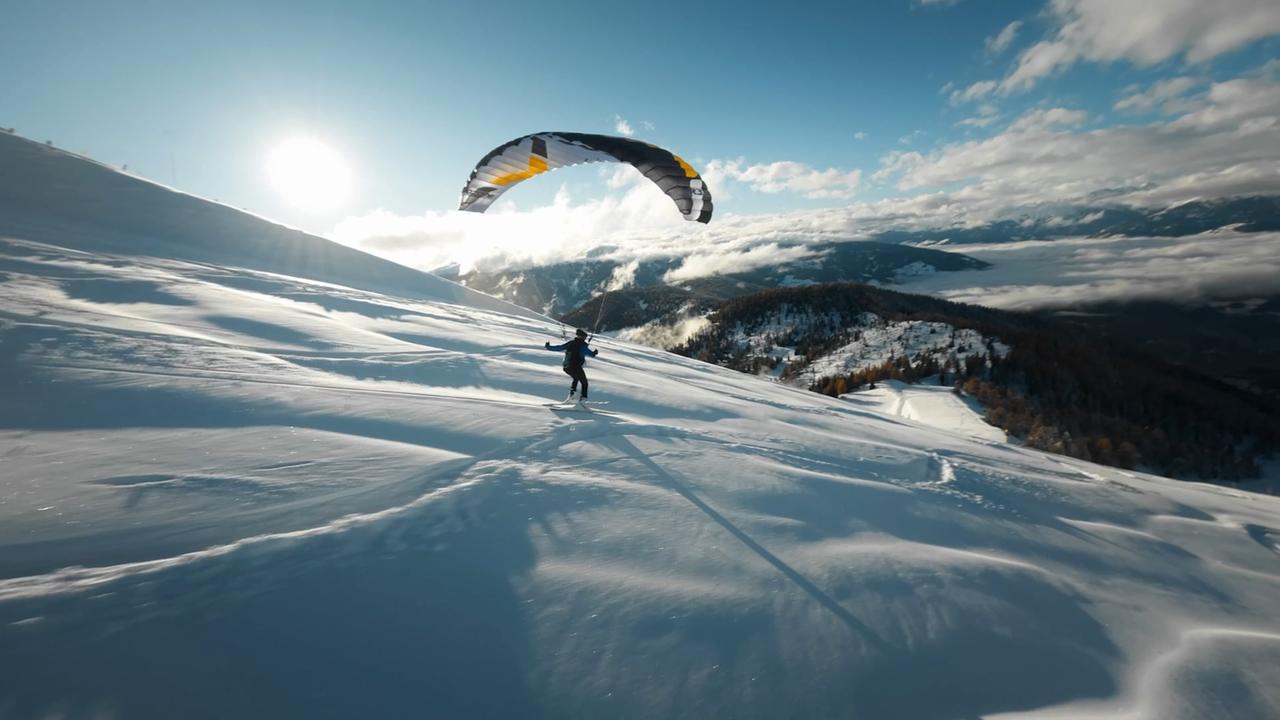 Das Bild zeigt eine schneebedeckte Berglandschaft in Südtirol. Im Vordergrund ist eine Person zu sehen, die mit Skiern und einem großen Gleitschirm, der sich über ihr ausbreitet, eine Abfahrt macht. Die Skifahrerin oder der Skifahrer steht auf einer schneebedeckten Fläche und hat die Arme ausgebreitet. Der Gleitschirm hat eine markante schwarz-gelbe Farbe und ist im Aufwind.   Im Hintergrund erstrecken sich hügelige Berge, einige mit Bäumen bepflanzt, während andere steil und schneebedeckt sind. Der Himmel ist blau mit vereinzelten Wolken und die Sonne scheint hell, was eine klare und lebendige Atmosphäre schafft. Die Szene vermittelt ein Gefühl von Freiheit und Abenteuer, typisch für moderne Outdoorsportarten. Die Beschriftung deutet darauf hin, dass es sich um Speedriding handelt, eine Aktivität, die Skifahren und Gleitschirmfliegen kombiniert.