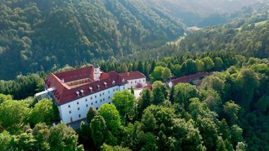 Das Bild zeigt eine malerische Landschaft im Sulmtal, Weststeiermark, Österreich. Im Vordergrund befindet sich ein großes, dreigeschossiges Gebäude mit weißen Wänden und roten Dächern. Das Gebäude ist umgeben von üppigem Grün; Bäume und Sträucher bilden eine dichte Vegetation. In der oberen rechten Bildhälfte sind sanfte Hügel zu sehen, die ebenfalls bewaldet sind.  Hinter dem Gebäude schlängelt sich ein Fluss durch das Tal. Die Landschaft ist von sanften Hügeln und bewaldeten Bereichen geprägt, was ein ruhiges und idyllisches Bild vermittelt. Die Lichtverhältnisse deuten auf einen sonnigen Tag hin, wobei die Sonne durch die Bäume scheint und das Bild aufhellt.   Das Bild könnte aus der ORF/3sat-Dokumentation über das Sulmtal stammen, die die Natur und Kultur der Region thematisiert.