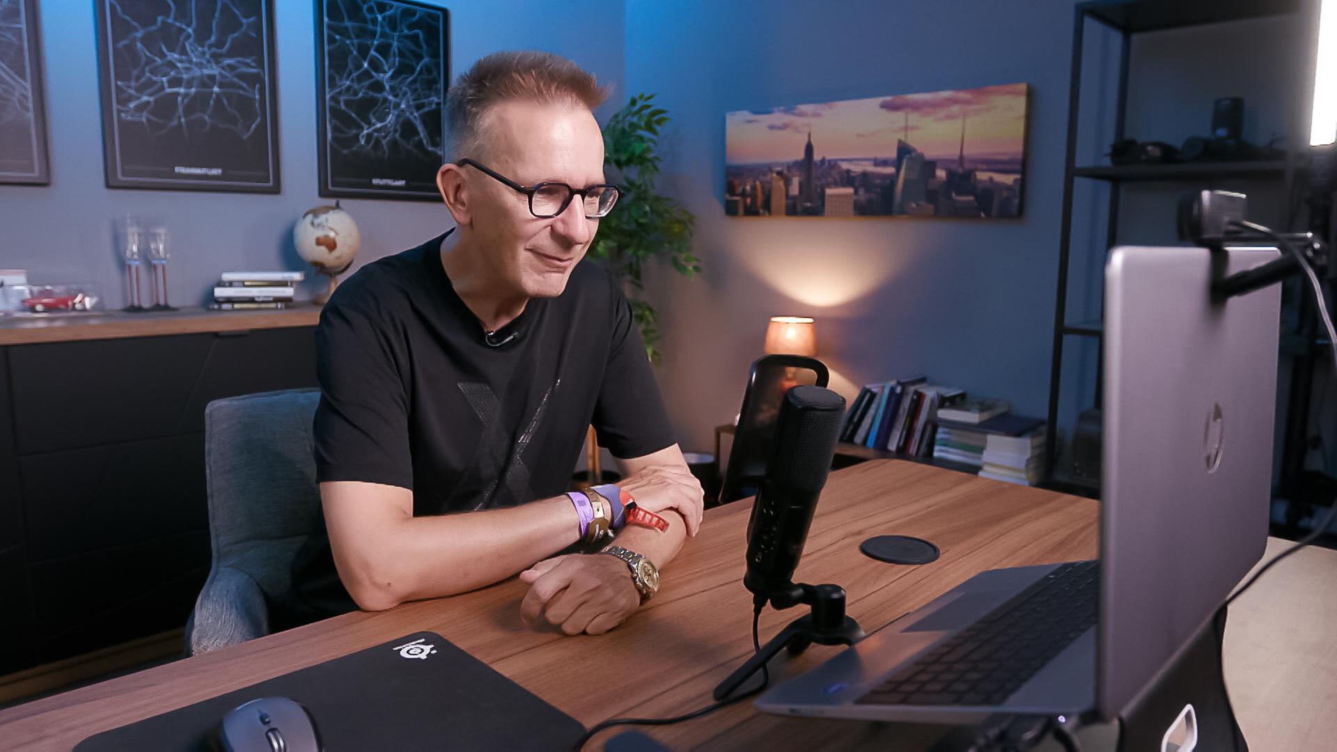 Das Bild zeigt einen Mann, der am Schreibtisch sitzt und an einem Computer arbeitet. Er trägt eine schwarze T-Shirt mit einem dezenten Muster und eine Brille mit dünnem Rahmen. Auf seinem Arm sind mehrere Armbänder sichtbar, und am Handgelenk trägt er eine Armbanduhr.  Der Schreibtisch ist aus Holz und darauf befindet sich ein Mikrofon, ein Laptop und ein schwarzes Mauspad. Im Hintergrund sind Regale mit Büchern und ein Globus zu sehen. An der Wand hängen zwei Poster mit abstrakten Karten von Städten (Frankfurt und Stuttgart). Neben steht eine Lampe, die eine angenehme Beleuchtung bietet, und an der Wand gegenüber ist ein Bild einer Skyline mit Hochhäusern in der Dämmerung.  Die gesamte Szene vermittelt eine moderne und professionelle Atmosphäre, die typisch für einen Arbeitsbereich ist.