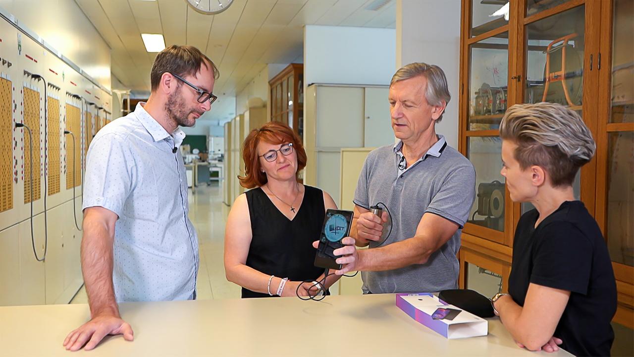 Das Bild zeigt vier Personen in einem Innenraum, der wie ein Labor oder ein technisches Studio aussieht, mit Regalen und Geräten im Hintergrund. Auf einem Tisch in der Mitte steht ein kleines, tragbares Gerät, das von einem Mann gehalten wird. Neben ihm stehen drei weitere Personen, die aufmerksam auf das Gerät schauen.   Die Personen sind folgendermaßen positioniert:  - Links steht ein Mann mit Brille, der ein Hemd mit Punkten trägt.  - In der Mitte steht eine Frau mit schulterlangen, lockigen roten Haaren, die ein schwarzes Oberteil trägt und das Gerät in der Hand hat.  - Neben ihr steht ein älterer Mann mit kurzen, grauen Haaren und einem hellen, kurzärmeligen Hemd. Er hält das Gerät und erklärt wahrscheinlich dessen Funktion. - Rechts sitzt eine weitere Frau mit kurzem, blonden Haar, die ein schwarzes T-Shirt trägt und ebenfalls auf das Gerät schaut.  Der Raum hat eine helle, neutrale Farbgebung, die durch die Regale und Wände verstärkt wird. An den Wänden sind technische Geräte und Ausrüstungen zu sehen, die auf eine wissenschaftliche Umgebung hinweisen.