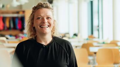 Eine lachende Frau mit lockigem blondem Haar steht in einem hellen Speisesaal mit leeren Tischen und Stühlen. Im Hintergrund hängen bunte Jacken an einer Garderobe.