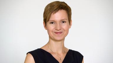 Eine Frau mit kurzen Haaren steht vor weißem Hintergrund und lächelt in die Kamera.