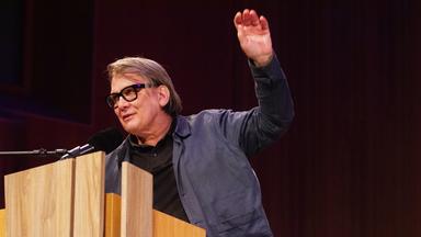 Ein Mann mit dicker schwarzer Brillenfassung spricht an einem Pult in ein Mikrofon und hebt die linke Hand.