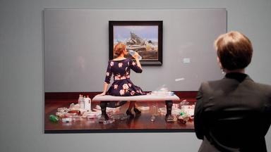 Eine Frau betrachtet ein großformatiges Foto an der Wand, das eine junge Frau zeigt, die vor einem Bild von Caspar David Friedrich im Museum sitzt und aus einer Plastikflasche trinkt. Um sie herum liegen leere Plastikverpackungen und Flaschen.