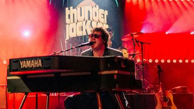 Eine Band auf der Bühne. Der Mann in der Mitte sitzt an einem Keyboard und singt.