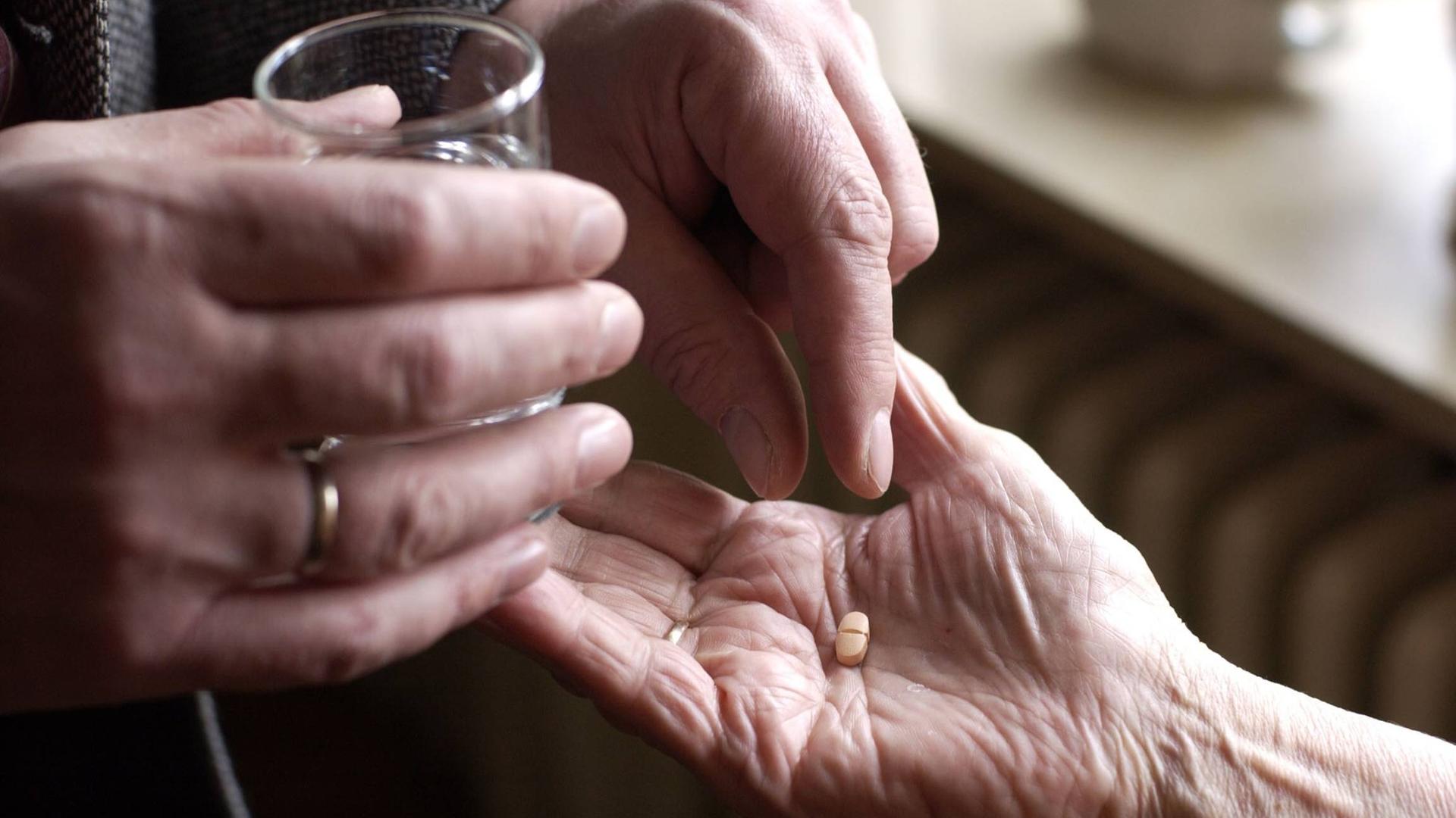 Auf dem Bild ist ein Paar Hände zu sehen. Eine Hand hält ein Wasserglas, während die andere eine Pille in die Hand einer alten Frau legt.