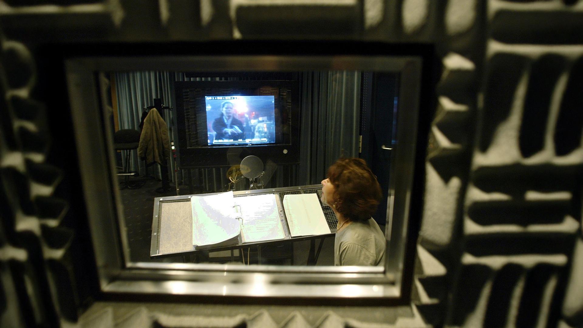 Eine Frau sitzt hinter einer Glasscheibe in einem kleinen Studio und spricht in ein Mikrofon, während sie auf einem Bildschirm einen Film schaut.