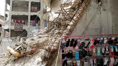 In der syrischen Stadt Douma steht zwischen Gebäudetrümmern  ein Verkaufsstand mit bunten Socken auf der Straße, im Hintergrund sieht man in einer Ruine bunte Kinderwäsche auf einem Seil zum Trocknen aufgehängt.