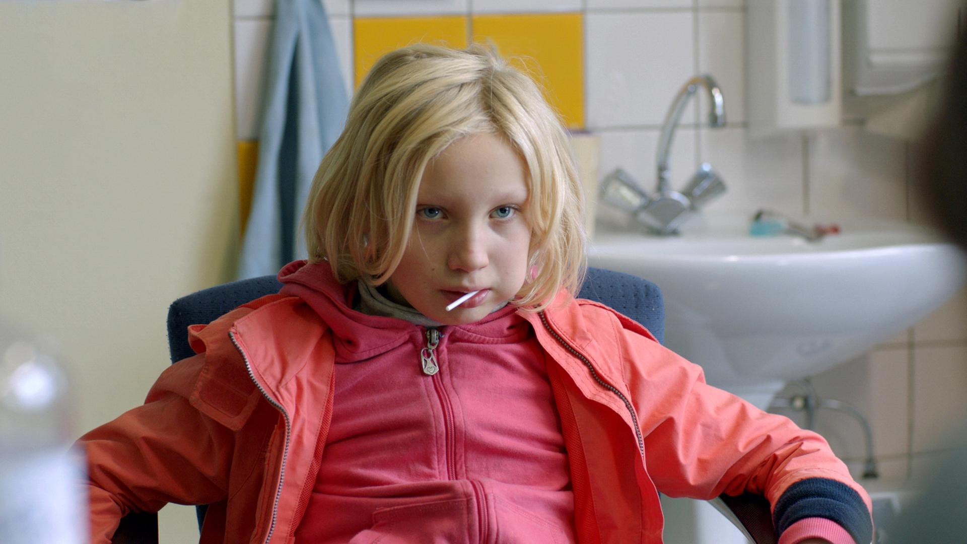 Benni (Helena Zengel), ein etwa 10-jähriges Mädchen in rosa Jacke und Anorak mit einem Lolly im Mund, lümmelt auf einem Stuhl und sieht ihr Gegenüber finster an. Im Hintergrund ein Waschbecken mit Seifen- und Papierspender an einer gekachelten Wand.