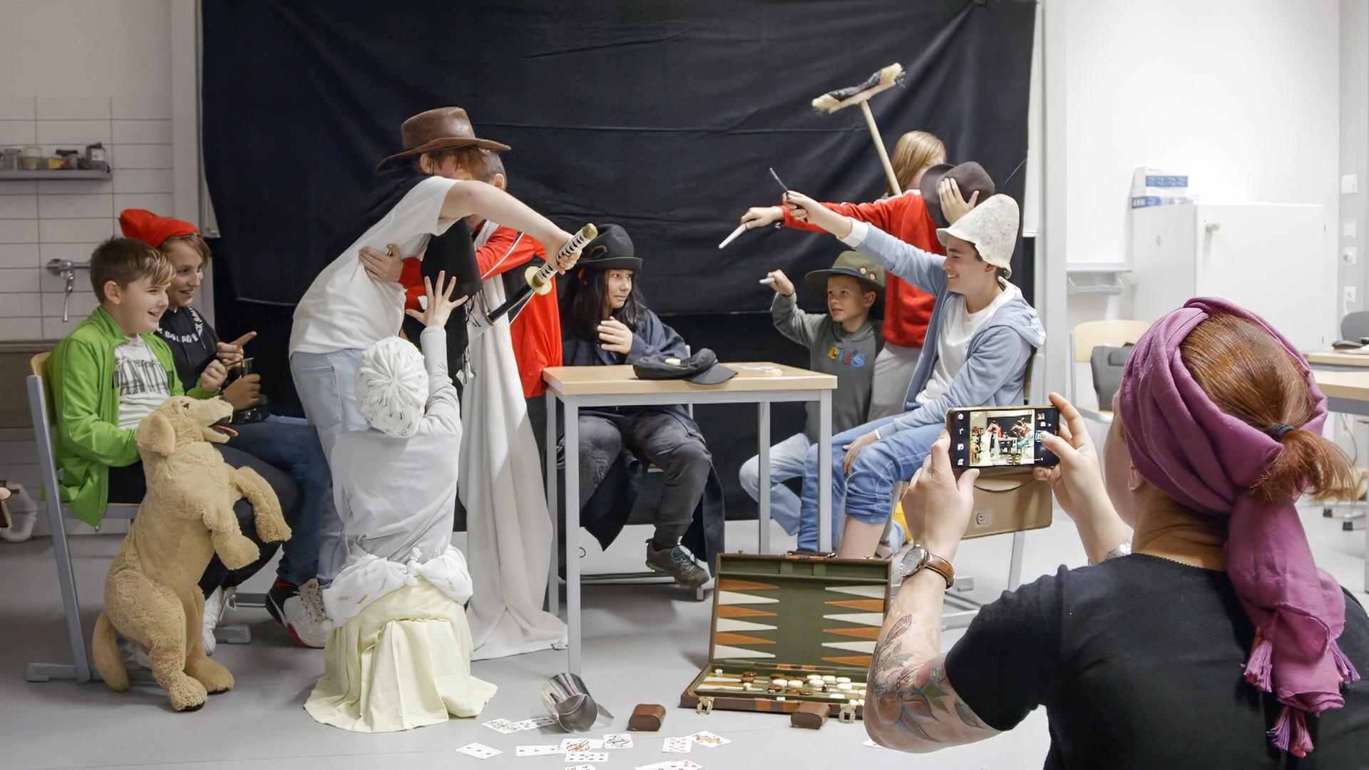 Eine Gruppe von Schülern in einem Klassenraum stellt ein lebendes Gemälde dar, während eine Person im Vordergrund ein Foto davon macht. Die Schüler tragen verschiedene Kostüme und Requisiten.