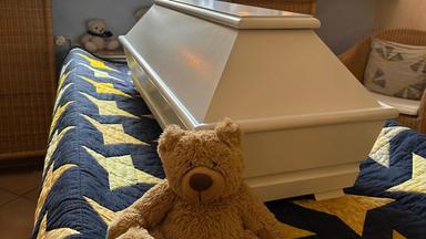 Heller Kindersarg auf einer Sternendecke, davor ein Teddy, im Hintergrund ein Buntglasfenster und brennende Windlichter.