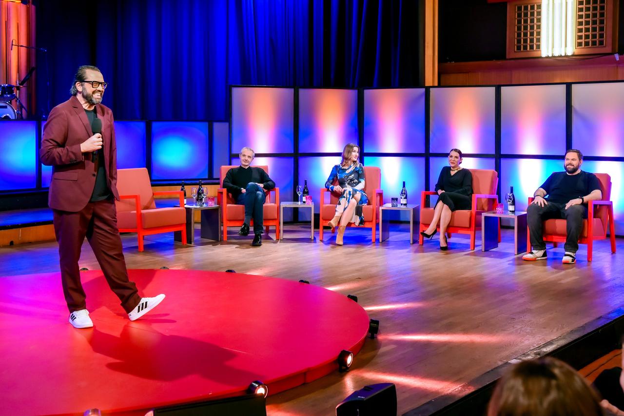 Das Bild zeigt eine Bühne, die für eine Veranstaltung des satirischen Monatsrückblicks "Tafelrunde" vorbereitet ist. Im Vordergrund steht ein rednerisches Podest in runder Form, das rot ist. Während eines Auftritts ist ein Mann in einem burgunderfarbenen Anzug mit einer schwarzen T-Shirt und weißen Sportschuhen zu sehen. Er hält ein Mikrofon und spricht in die Richtung des Publikums.  Hinter ihm sitzen fünf Personen auf rotfarbenen Stühlen, die in einer Halbkreisformation angeordnet sind. Diese Personen blicken auf ihn. Auf den Tischen vor ihnen stehen Gläser und Weinflaschen. Die Frauen tragen elegante Outfits: Eine Frau in einem bunten Kleid mit blauen und weißen Mustern, eine andere in einem schwarzen, langen Kleid. Die Herren tragen mostly schwarze und dunkelblaue Kleidung.   Die Beleuchtung der Bühne ist in verschiedenen Farben gehalten, überwiegend in Blau- und Orangetönen, was eine auffällige Atmosphäre schafft. Im Hintergrund sind einige Musikinstrumente vermutlich für eine Live-Performance zu sehen. Die gesamte Szene strahlt eine unterhaltsame und lebendige Stimmung aus.