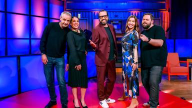 Die Aufnahme zeigt fünf Personen, die auf einer Bühne stehen. Der Boden der Bühne ist rot, und im Hintergrund ist eine helle Kulisse mit bunten Lichtern in Blau und Violett.   Von links nach rechts: 1. Ein Mann mit grauen Haaren, trägt ein schwarzes Sweatshirt und eine dunkle Jeans. Er hat einen leicht scherzhaften Ausdruck. 2. Eine Frau in einem schwarzen Kleid mit Falten und einem eleganten Oberteil. Ihr Gesichtsausdruck ist freundlich. 3. Der dritte Mann trägt einen burgunderfarbenen Anzug und eine schwarze T-Shirt darunter, mit einer offenen Körperhaltung und einem einladenden Lächeln. 4. Eine weitere Frau, die ein Kleid mit blauen und schwarzen Farbtönen trägt. Sie steht mit einem Lächeln und gestreckten Armen da. 5. Der letzte Mann trägt ein schwarzes T-Shirt und eine dunkle Jeans, schaut in die Kamera und deutet mit dem Finger auf etwas im Bild.  Im Hintergrund sind einige Musikinstrumente zu erkennen, und auf der Bühne steht ein rotes Sofa. Die Szenerie hat eine lebendige, unterhaltsame Atmosphäre.