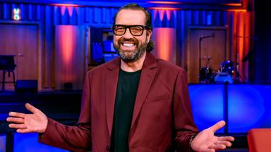 Das Bild zeigt einen Mann, der in einem Raum mit einer Bühne und farbigen Lichtern steht. Er trägt einen rotbraunen Blazer über einem schwarzen T-Shirt. Der Mann hat einen kurzen, gepflegten Bart und trägt eine Brille. Er macht eine offene Handbewegung mit seinen Armen, was Freundlichkeit und Offenheit ausdrückt. Im Hintergrund sind unscharf einige Instrumente und Bühnenausstattungen zu erkennen, die auf eine Unterhaltungssituation hindeuten. Die Atmosphäre wirkt lebhaft und einladend.