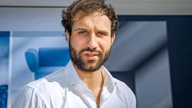 Porträtbild eines jungen Mannes mit Bart und in einem weißen Hemd mit leger offenem Kragen.