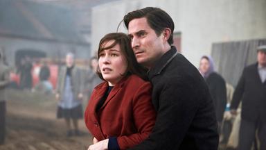 "TANNBACH - Schicksal eines Dorfes : Schatten des Krieges": Pastor Wolfgang Herder (Clemens Schick) hat seine Arme um Annas (Henriette Confurius) Taille gelegt und drückt sie fest an sich. Sie schauen in die gleiche Richtung.