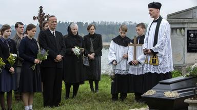 Trauergemeinde bei einer Beerdigung auf einem Friedhof; ein Priester und Ministranten stehen neben einem schwarzen Sarg, während Angehörige mit Blumen in der Hand Abschied nehmen.