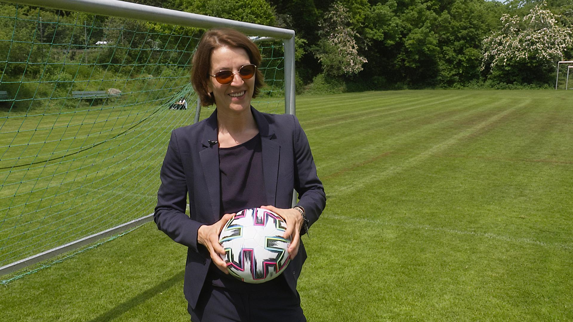 Eine Frau im dunklen Hosenanzug steht mit einem Fußball in der Hand vor einem Tor auf einem Fußballplatz im Grünen und lächelt in die Kamera.