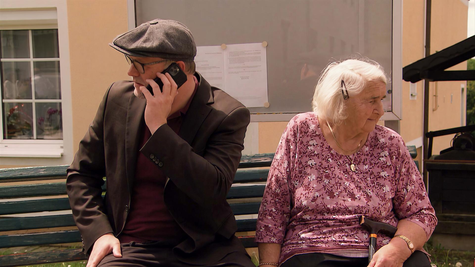 Das Bild zeigt eine Szene, die während der Dreharbeiten zu einer TV-Dokumentation stattfindet. Im Vordergrund sitzen zwei Personen auf einer Bank. Auf der linken Seite ist ein Mann zu sehen, der ein Handy in der Hand hält und telefoniert. Er trägt eine schwarze Jacke und einen grauen Hut. Auf der rechten Seite sitzt eine ältere Frau mit schneeweißem Haar, die ein rosa Shirt trägt und einen Gehstock dabei hat. Beide blicken in unterschiedliche Richtungen. Hinter ihnen ist eine Wand mit einem Fenster und einem Plakat oder einer Mitteilung, die an einem Brett befestigt ist. Die Umgebung vermittelt einen ländlichen Charakter, wahrscheinlich in einer idyllischen Gemeinde im Südburgenland.