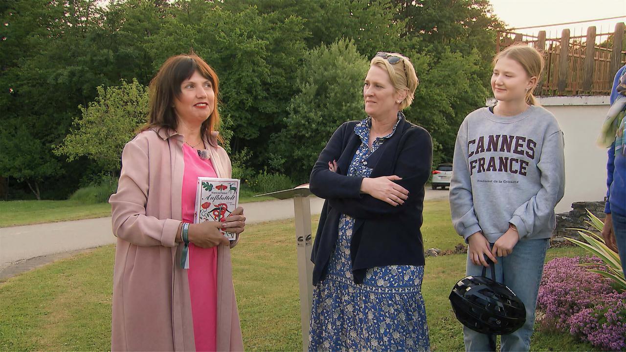 Das Bild zeigt eine Szene im Freien, wo mehrere Personen zusammenstehen. Im Vordergrund steht eine Frau mit langen, braunen Haaren, die ein pinkes Kleid und eine beige Jacke trägt. Sie hält ein Buch in der Hand. Neben ihr steht eine weitere Frau mit kurzen, blonden Haaren, die eine dunkle Jacke über einem geblümten Kleid trägt. Die dritte Person ist ein Mädchen, das einen grauen Sweater mit der Aufschrift "CANNES FRANCE" und Jeans trägt, und eine Fahrradhelm in der Hand hält.   Im Hintergrund ist eine grüne Landschaft mit Bäumen zu sehen. An der linken Bildseite führt eine asphaltierte Straße vorbei. Rechts im Bild sind fliederfarbene Blumen zu erkennen. Es scheint später Nachmittag oder Abend zu sein, da das Licht weich ist. Die Personen stehen entspannt und lächeln.