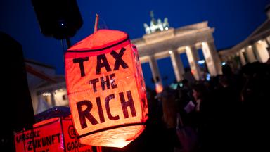 Bei einer Demo vor dem Brandenburger Tor bei Nacht ist im Vordergrund eine rote Lampe mit dem Text tax the rich zu sehen.