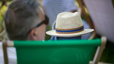 Ein Mann mit grauen Haaren sitzt in einem grünen Liegestuhl und hält einen Strohhut mit blau-braunem Band auf dem Schoß; die Szene spielt sich im Garten ab.