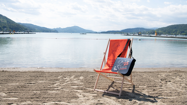 Liegestuhl und Tasche mit Bachmannpreis-Logo am Ufer eines Sees mit Bergen im Hintergrund