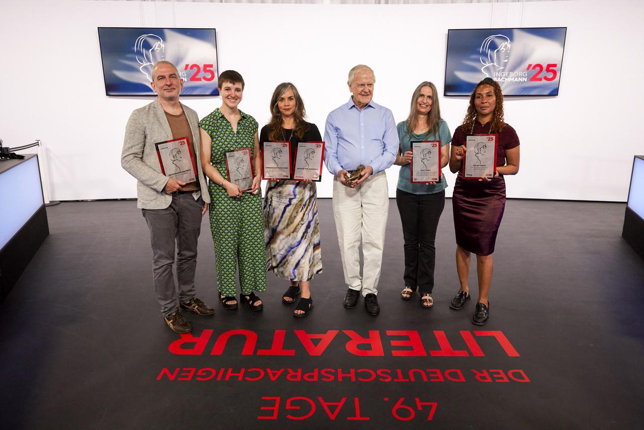 Gruppenfoto der Preisträger:innen der 49. Tage der deutschsprachigen Literatur 2025 mit ihren Auszeichnungen, aufgenommen im Studio vor zwei Bildschirmen mit der Aufschrift Ingeborg Bachmann '25.