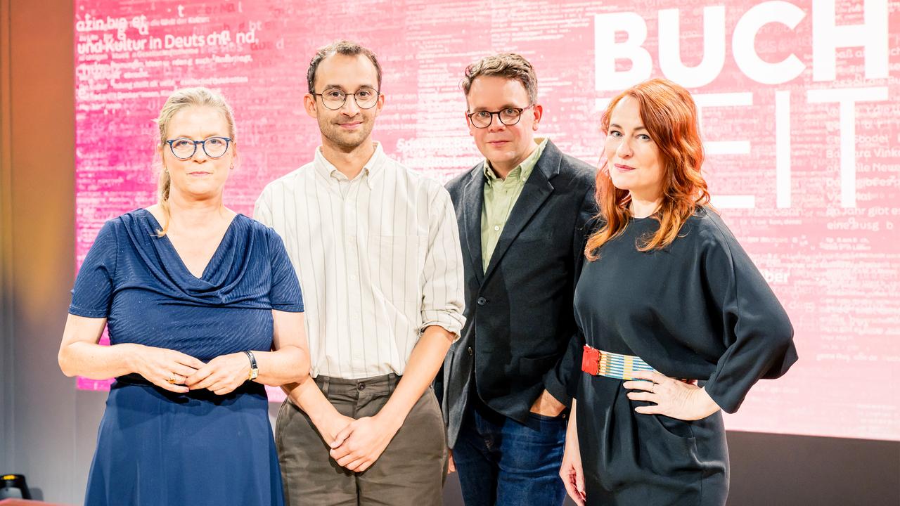 Das Foto zeigt das Team der Sendung „Buchzeit“ auf der Frankfurter Buchmesse 2025. Vier Personen stehen nebeneinander vor einem pink-roten Hintergrund mit weißem Schriftzug, in dem das Wort „BUCHZEIT“ deutlich zu erkennen ist. Zwei der Personen tragen Brillen. Die Kleidung reicht von leger bis semi-formell. Alle blicken direkt in die Kamera.