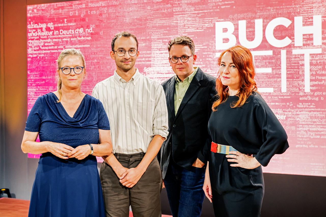 Das Foto zeigt das Team der Sendung „Buchzeit“ auf der Frankfurter Buchmesse 2025. Vier Personen stehen nebeneinander vor einem pink-roten Hintergrund mit weißem Schriftzug, in dem das Wort „BUCHZEIT“ deutlich zu erkennen ist. Zwei der Personen tragen Brillen. Die Kleidung reicht von leger bis semi-formell. Alle blicken direkt in die Kamera.