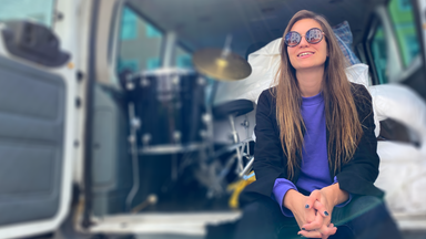 Junge Frau mit Sonnenbrille sitzt lächelnd auf der Ladefläche eines Kleintransporters, im Hintergrund ist ein Schlagzeug zu erkennen.